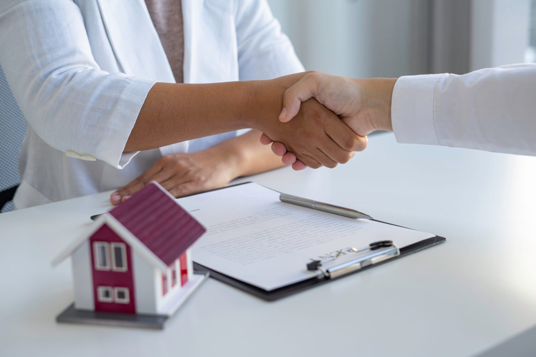 an image of two people shaking hands with a contract and model house in front of them to denote a story about residential transactions