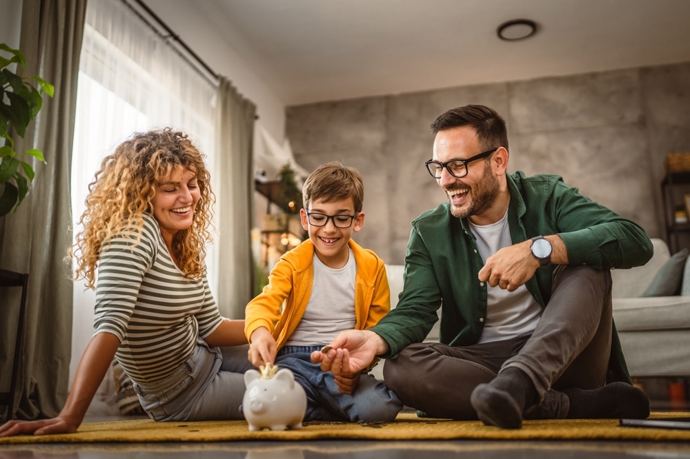 a family on the floor while a child puts a penny into a piggy bank to denote a blog on financial education